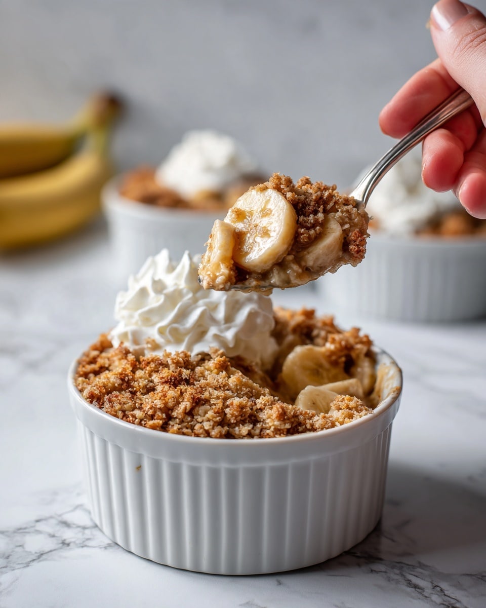 The image shows a close-up of a white round baking dish filled with a banana crumb casserole. The top layer is golden brown with a crumbly texture, dotted with small pieces of baked crumble. On top, several banana slices add a light yellow color and smooth texture. A spoon lifts a scoop of the casserole, revealing moist banana and crumble layers inside, with a dollop of white cream on the side. The background is a white marbled surface, and a blue cloth is partly visible beneath the dish. Photo taken with an iphone --ar 4:5 --v 7