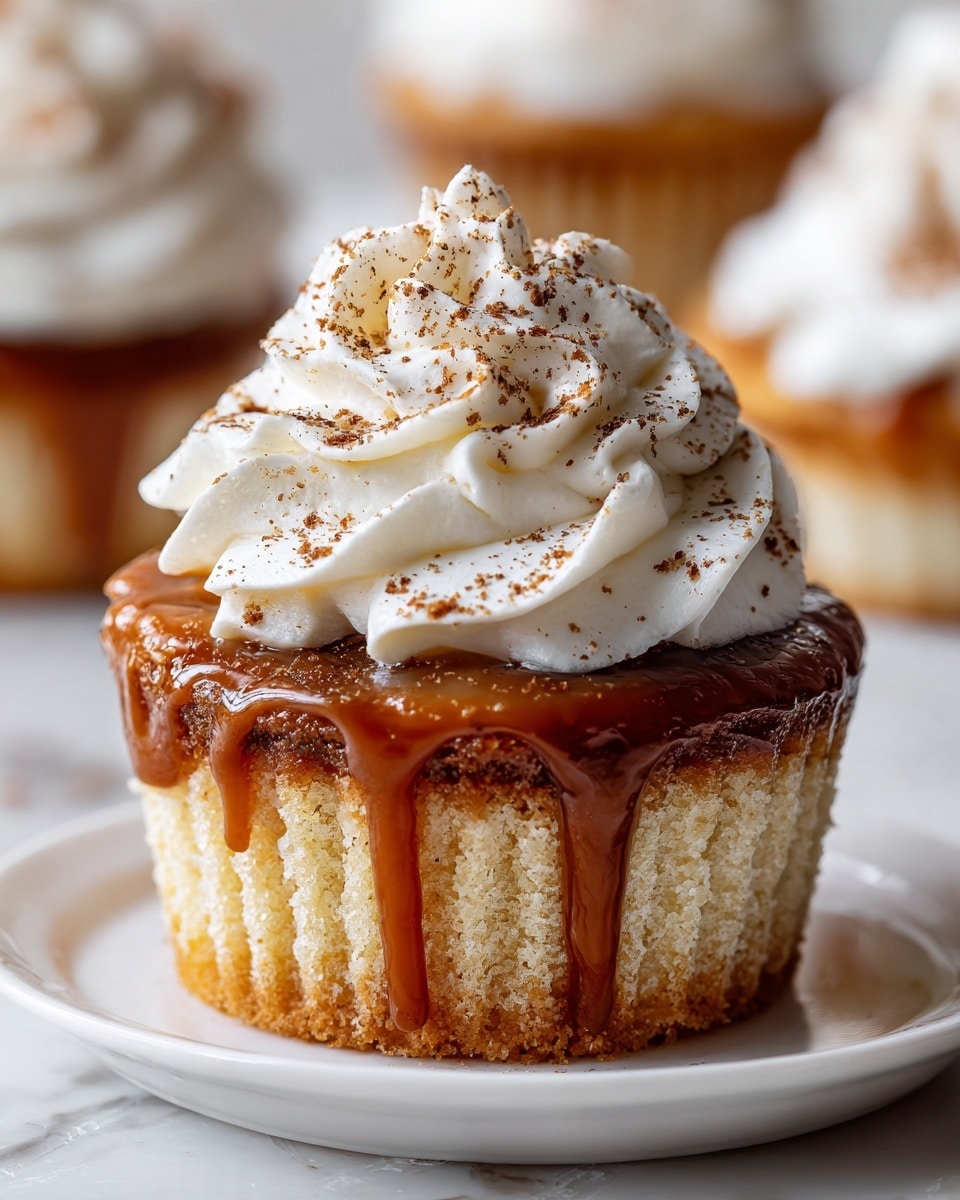 A small cupcake with a golden brown base topped with a thick layer of smooth caramel sauce that drips over the side, crowned by a swirl of fluffy white whipped cream sprinkled lightly with brown spice powder; the cupcake is placed on a white plate with a slightly shiny finish, set on a white marbled surface with other similar cupcakes softly blurred in the background. photo taken with an iphone --ar 4:5 --v 7