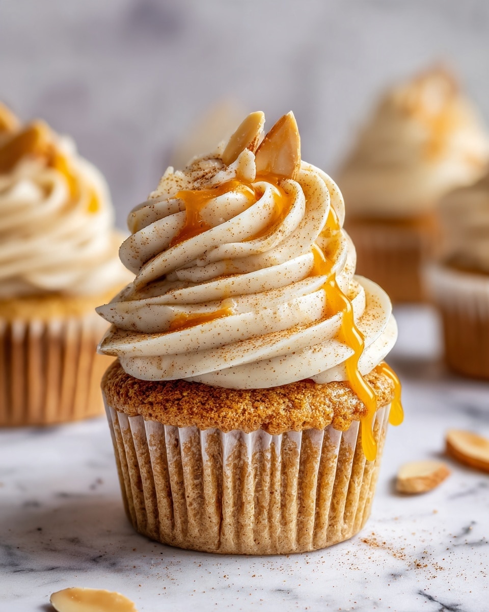 A single vanilla cupcake with a light golden base rests on a white marbled surface, topped with a swirl of creamy beige frosting layered in thick, soft waves about three layers high. The frosting is drizzled with a shiny, amber-colored caramel sauce that glistens and flows down the sides slightly. Thin, pale almond slices embellish the frosting, with one prominent almond slice placed upright at the top, adding texture contrast. Other similar cupcakes are softly blurred in the background, also decorated with the frosting, sauce, and almond slices. photo taken with an iphone --ar 4:5 --v 7