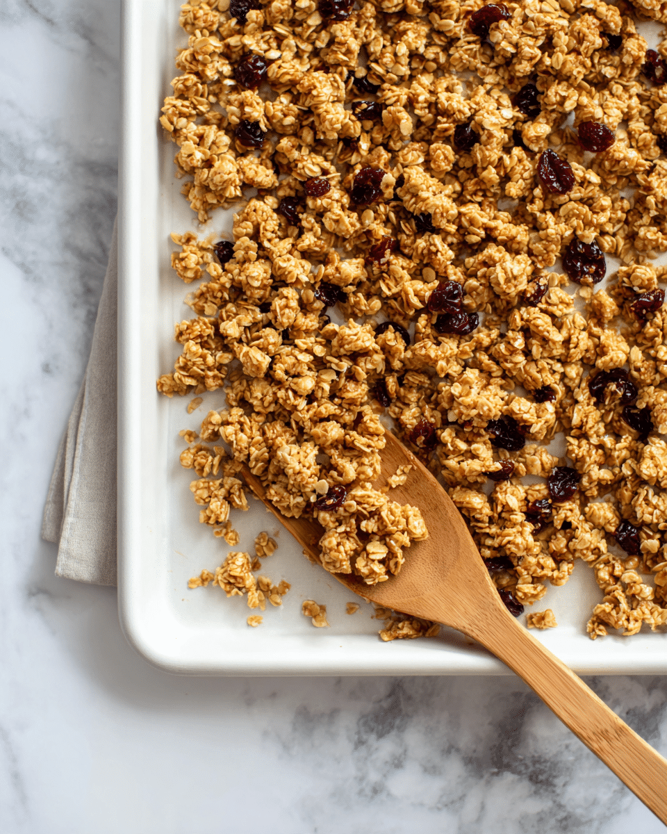 A large white baking tray filled with golden brown oat clusters mixed with small dark red berries, spread unevenly with some clusters forming small mounds and others scattered. The oat clusters have a rough and crumbly texture, showing bits of oats sticking out. A wooden spatula lies on the right side, partially scooping some of the clusters. The tray rests on a white marbled surface, adding a clean, bright background. photo taken with an iphone --ar 4:5 --v 7