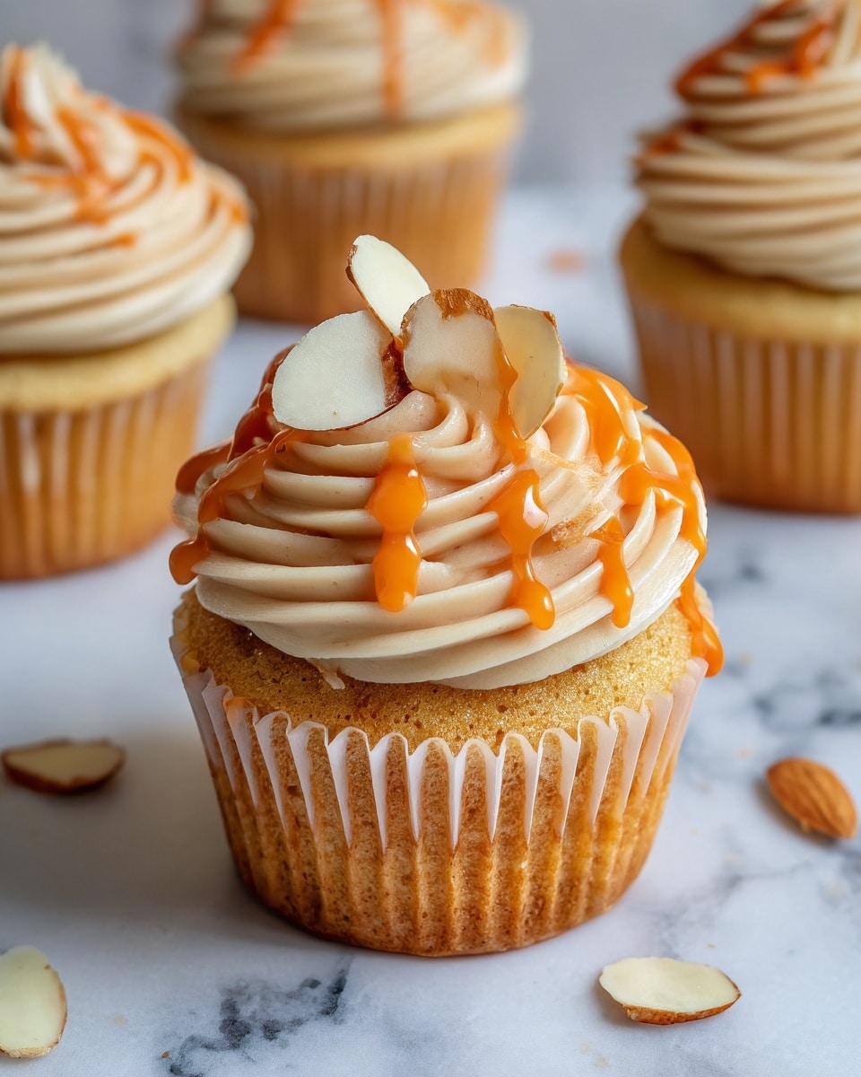 A single cupcake with a light golden base topped with two large swirls of creamy beige frosting. The frosting is smooth and thick with a shiny orange drizzle cascading down its layers and the sides of the cupcake. On top, there are three thin almond slices arranged with one standing upright and two lying flat. The cupcake sits on a white marbled surface, surrounded by similar cupcakes in the blurred background. photo taken with an iphone --ar 4:5 --v 7